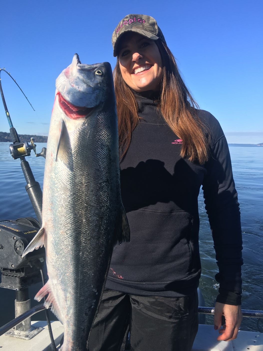 Woman smiling while holding a coho salmon with a fishing rod bent in the background.